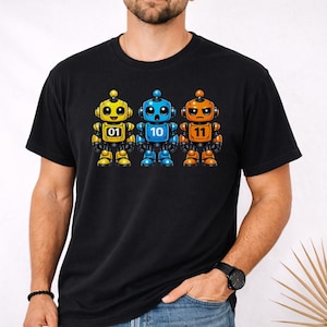 Op de afbeelding: Zwart t-shirt met drie cartoonrobots in geel, blauw en oranje. Elke robot heeft een nummer: 01, 10 en 11. De robots hebben ronde hoofden, antennes en gedetailleerde lichaamsontwerpen. Het shirt is een casual kledingstuk.