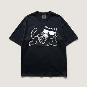 以下が含まれることがあります： サングラスをかけた白い漫画の猫がビールジョッキを持っている黒いTシャツ。 猫はくつろいでおり、ビールジョッキを手に持っています。 Tシャツは半袖で、クルーネックです。