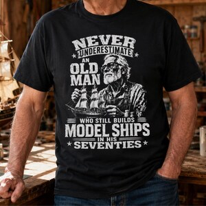 May include: A black short-sleeved t-shirt featuring a white graphic of an older person with glasses building a model ship. The text reads: NEVER UNDERESTIMATE AN OLD MAN WHO STILL BUILDS MODEL SHIPS IN HIS SEVENTIES, arranged in a bold, distressed font.