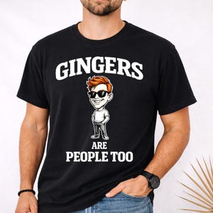 May include: Black crew neck t-shirt featuring the text "GINGERS" above a cartoon illustration of a redhead wearing sunglasses. Below the illustration, the text reads "ARE PEOPLE TOO".