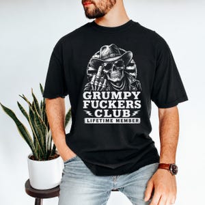 May include: Black t-shirt featuring a skull wearing a cowboy hat and leather jacket, giving the middle finger. The text "GRUMPY FUCKERS CLUB" and "LIFETIME MEMBER" are printed in white. The shirt is a casual style.