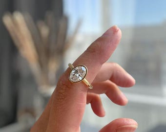 May include: A gold ring featuring an oval-cut diamond. The diamond is set in a bezel setting, and the band is a thin, polished gold. The ring is displayed on a finger, with a blurred background.