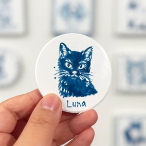 Hand holding a blue-and-white round cat portrait magnet labeled Luna, with other ceramic pet magnets softly blurred in the background.