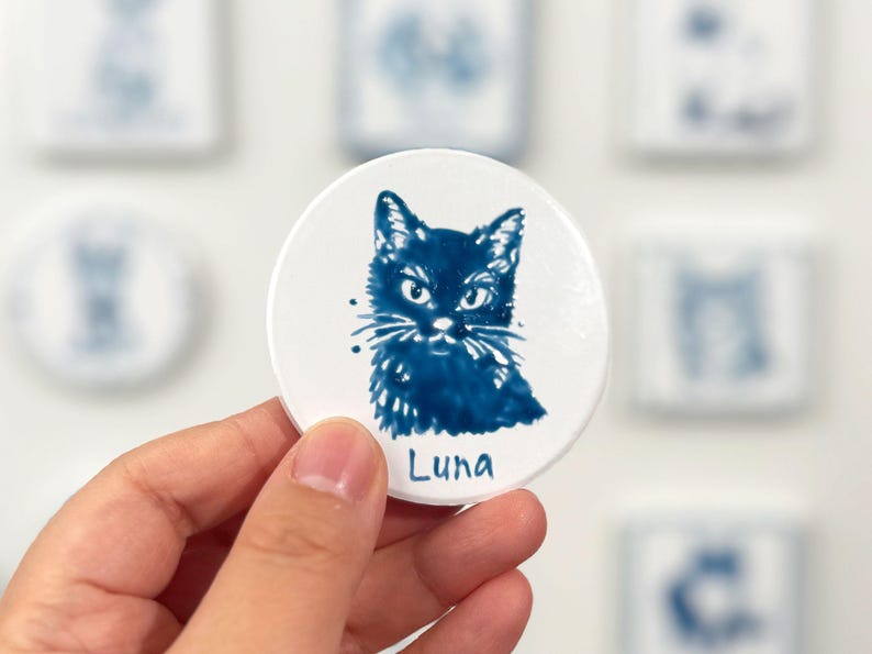 Hand holding a blue-and-white round cat portrait magnet labeled Luna, with other ceramic pet magnets softly blurred in the background.