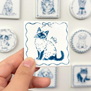 Hand holding a blue-and-white square Siamese cat portrait magnet labeled Miu, with matching custom pet magnets displayed behind it.