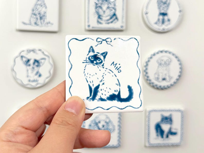 Hand holding a blue-and-white square Siamese cat portrait magnet labeled Miu, with matching custom pet magnets displayed behind it.