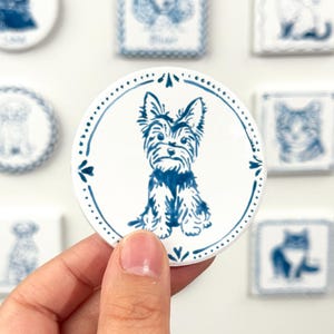 Hand holding a blue-and-white round Yorkshire Terrier portrait magnet against a softly blurred background of custom pet magnets.