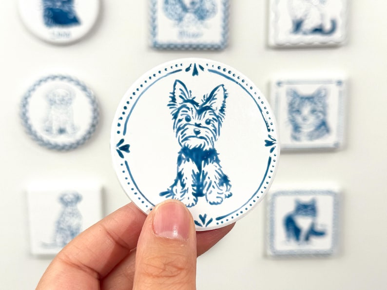 Hand holding a blue-and-white round Yorkshire Terrier portrait magnet against a softly blurred background of custom pet magnets.