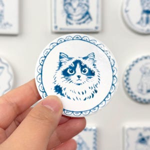 Hand holding a blue-and-white round ceramic magnet featuring a fluffy cat portrait, with other custom pet magnets softly blurred in the background.