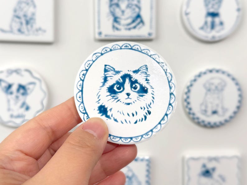 Hand holding a blue-and-white round ceramic magnet featuring a fluffy cat portrait, with other custom pet magnets softly blurred in the background.