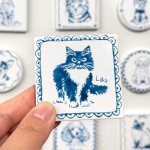 Hand holding a blue-and-white square ceramic magnet featuring a fluffy black cat portrait labeled Leo, with matching pet magnets blurred in the background.