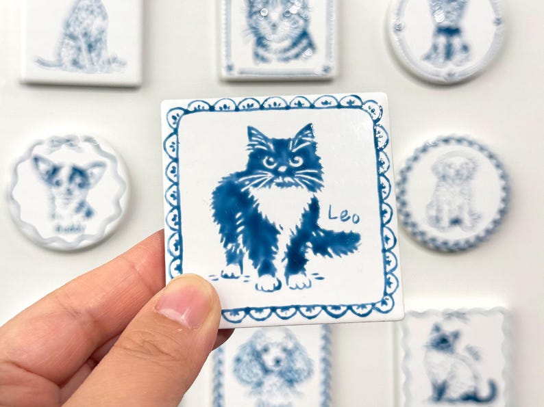 Hand holding a blue-and-white square ceramic magnet featuring a fluffy black cat portrait labeled Leo, with matching pet magnets blurred in the background.