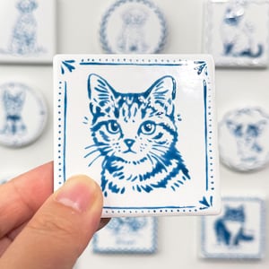 Hand holding a blue-and-white square cat portrait magnet against a softly blurred background of matching custom pet magnets.