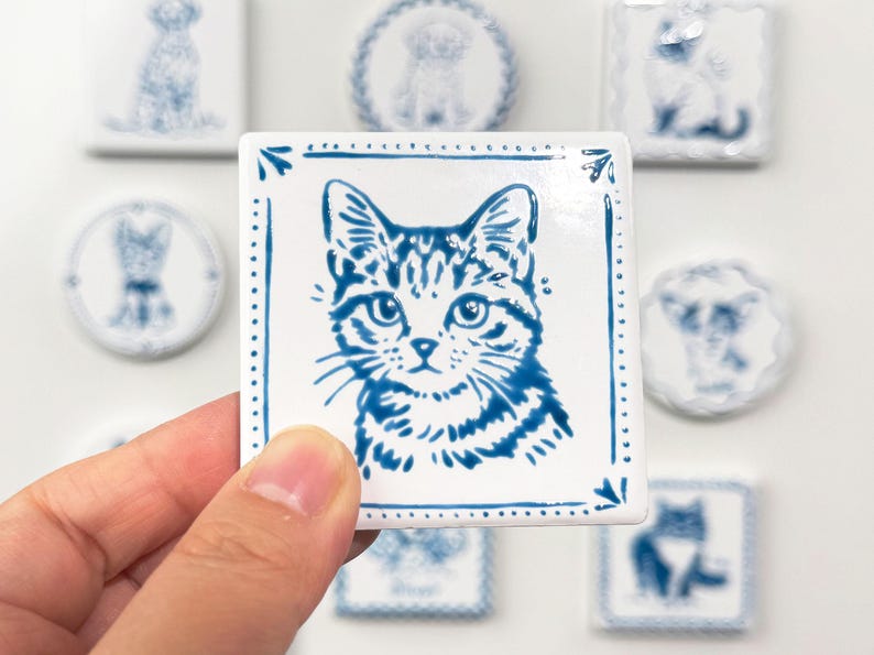 Hand holding a blue-and-white square cat portrait magnet against a softly blurred background of matching custom pet magnets.