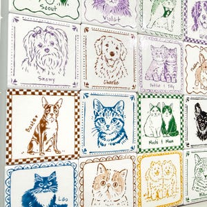 May include: A grid of square ceramic tiles featuring hand-drawn illustrations of various cats and dogs. Each tile displays a pet portrait in a distinct colour, accompanied by the animal's name written in cursive or print text below the illustration.