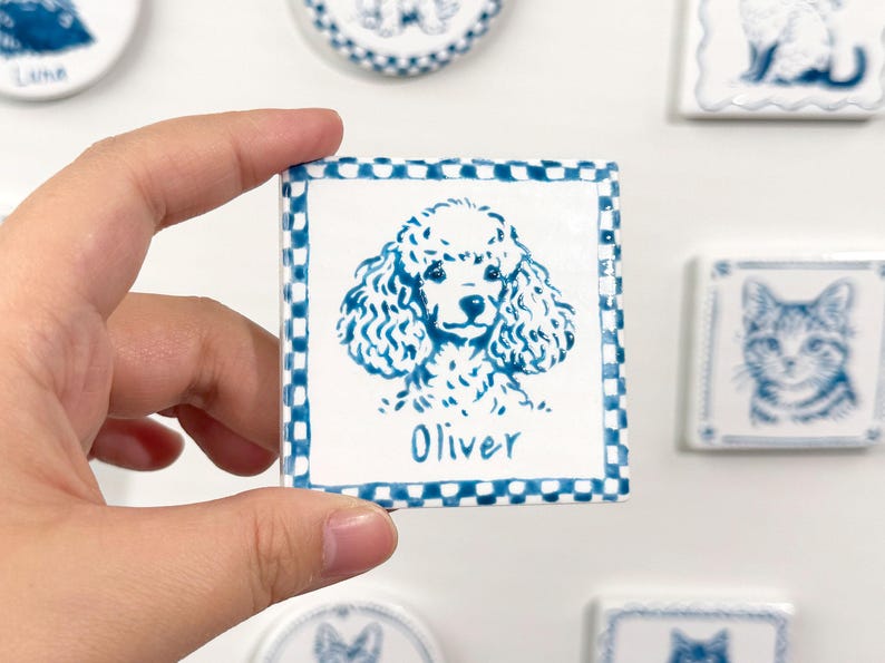 Hand holding a blue-and-white square poodle portrait magnet with the name Oliver, with other ceramic pet magnets blurred in the background.