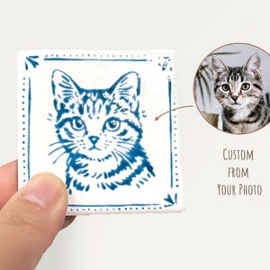 Hand holding a blue-and-white square cat portrait magnet next to a reference pet photo, showing a custom design made from the customer’s picture.