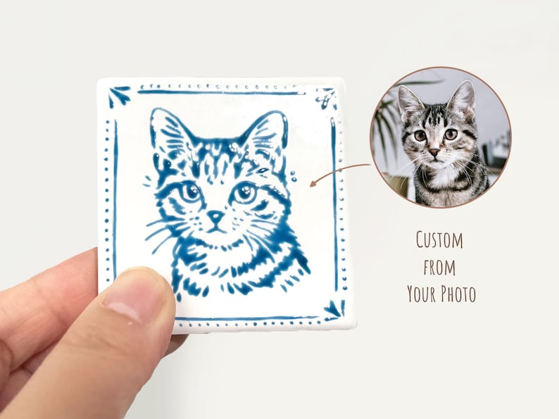 Hand holding a blue-and-white square cat portrait magnet next to a reference pet photo, showing a custom design made from the customer’s picture.