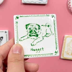 May include: A square, white ceramic tile with a green border and a green illustration of a pug wearing a bow tie. The name "Nugget" is written below the pug. Other dog-themed tiles are in the background.