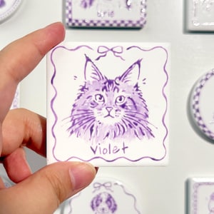 Op de afbeelding: Een vierkante witte keramische tegel met een paarse inktillustratie van een kattengezicht. De kat staat centraal met een klein strikje erboven getekend. De naam Violet staat in paars handschrift onder de kat. De tegel heeft een golvende rand.