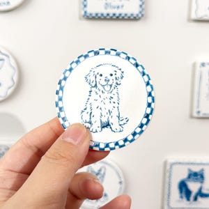 Hand holding a blue-and-white round dog portrait magnet with a checkered border against a blurred display of custom ceramic pet magnets.
