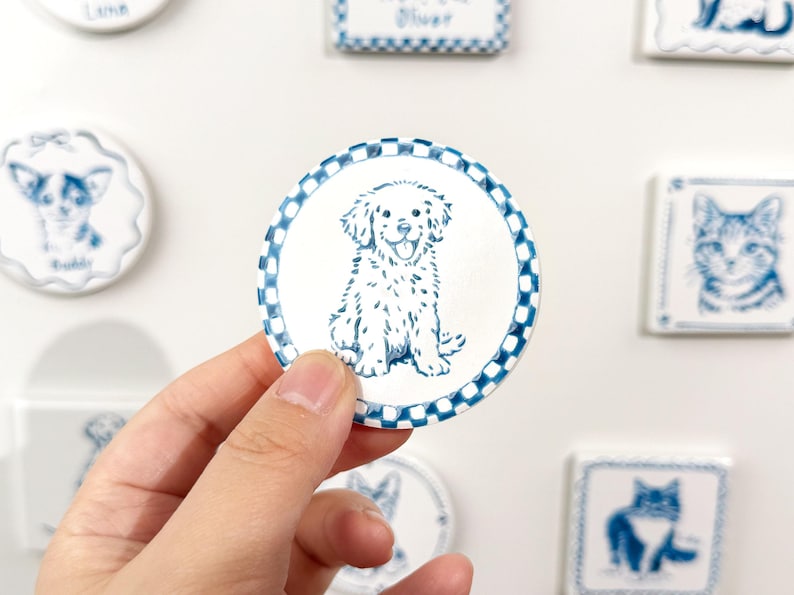 Hand holding a blue-and-white round dog portrait magnet with a checkered border against a blurred display of custom ceramic pet magnets.