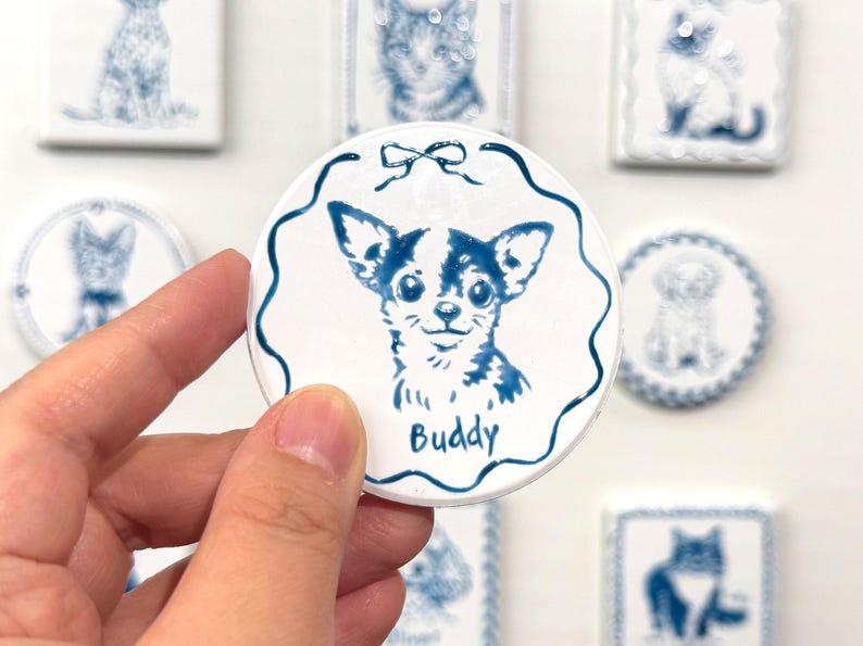 Hand holding a blue-and-white round Chihuahua portrait magnet labeled Buddy, with matching pet magnets displayed in the background.