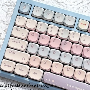 Może przedstawiać: Klawiatura w pastelowych kolorach z jasnoniebieską ramką i klawiszami w odcieniach różu, bieli i szarości. Klawiatura zawiera tekst "Peaceful Buddha Design Keyboard Keycaps" i "Eat a happy candy life".