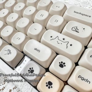 May include: Close-up of a white keyboard with custom keycaps. Some keycaps feature black paw prints, a cat face, and other designs. The word "shift" is printed on one keycap. The text "Peaceful Buddha Design Keyboard Keycaps" is visible.