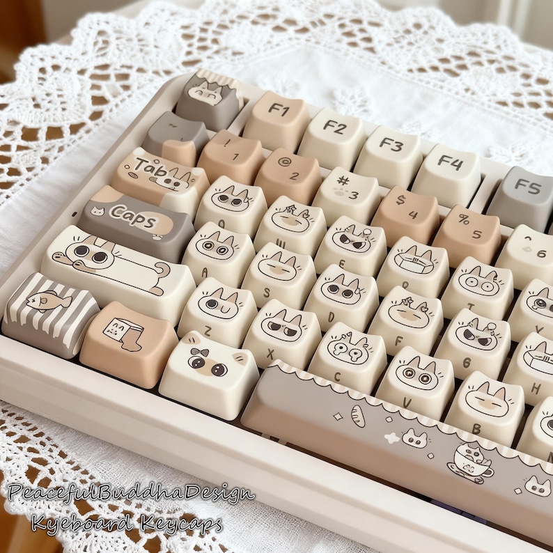 May include: A keyboard with custom keycaps featuring cat-themed designs in beige, brown, and white. The keycaps have various cat illustrations and text labels. The keyboard is set against a white background with a lace pattern.