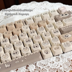 May include: A keyboard with custom keycaps featuring cat-themed designs. The keycaps are primarily off-white with black illustrations of cats with various expressions. The keyboard also has neutral-toned keys and the text "Peaceful Buddha Design Keyboard Keycaps".