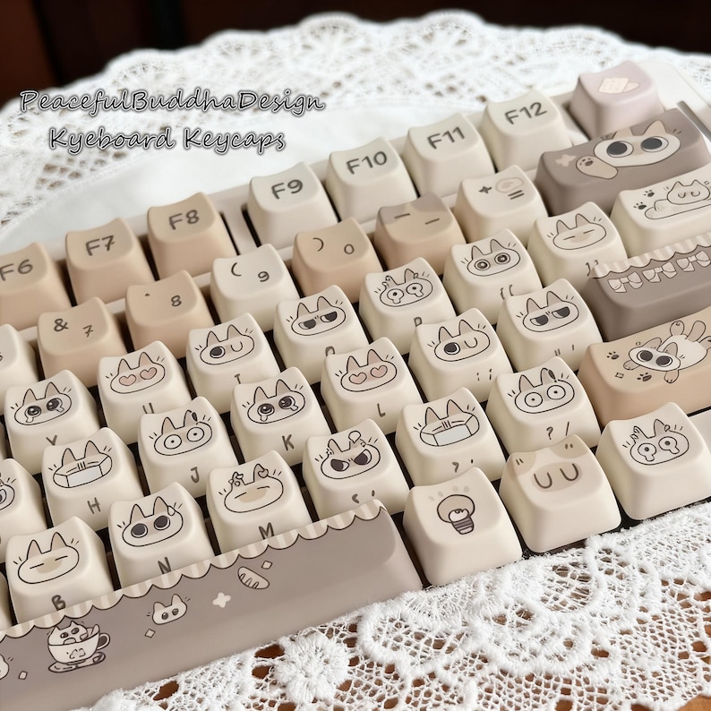 May include: A keyboard with custom keycaps featuring cat-themed designs. The keycaps are primarily off-white with black illustrations of cats with various expressions. The keyboard also has neutral-toned keys and the text "Peaceful Buddha Design Keyboard Keycaps".