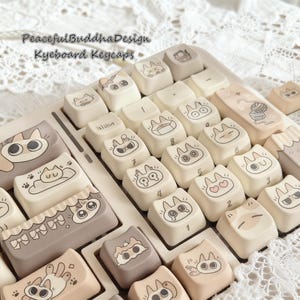 May include: A collection of off-white keyboard keycaps with cat-themed illustrations. The keycaps feature various cat designs, including faces, paws, and other cat-related graphics. The text "PeacefulBuddhaDesign Keyboard Keycaps" is visible.