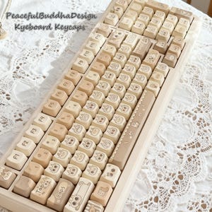 May include: A beige mechanical keyboard with custom keycaps. The keycaps feature a variety of cartoon character designs in black ink. The keyboard has a light beige frame and the text "PeacefulBuddhaDesign Keyboard Keycaps" is visible.
