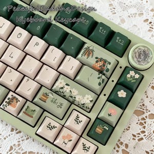 Puede incluir: Un teclado personalizado con teclas verde claro y teclas con ilustraciones botánicas. Las teclas tienen una variedad de diseños, incluyendo plantas en macetas y patrones florales.