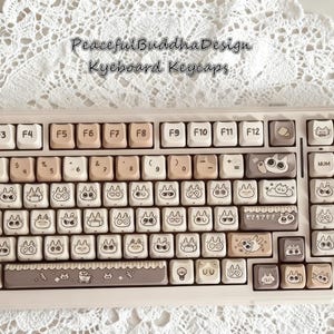 May include: A keyboard with custom keycaps featuring a light beige and brown color scheme. The keycaps have cute cartoon cat illustrations and the text "Peaceful Buddha Design Keyboard Keycaps".