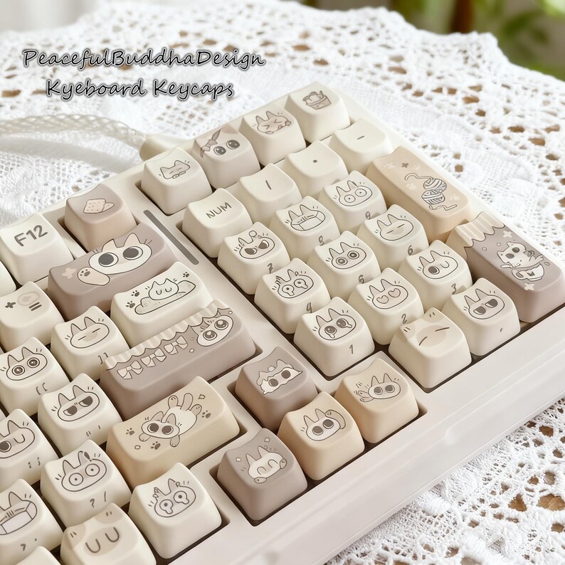May include: A keyboard with custom keycaps featuring cat illustrations. The keycaps are a mix of beige and taupe colors, with black line art drawings of cats in various poses. The keyboard has the text "Peaceful Buddha Design Keyboard Keycaps" at the top.