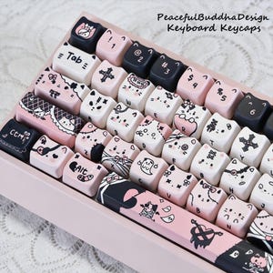 May include: A custom keyboard with pink and black keycaps. The keycaps feature cat-themed illustrations and symbols. The keyboard has the text "PeacefulBuddhaDesign Keyboard Keycaps" in the upper right corner.