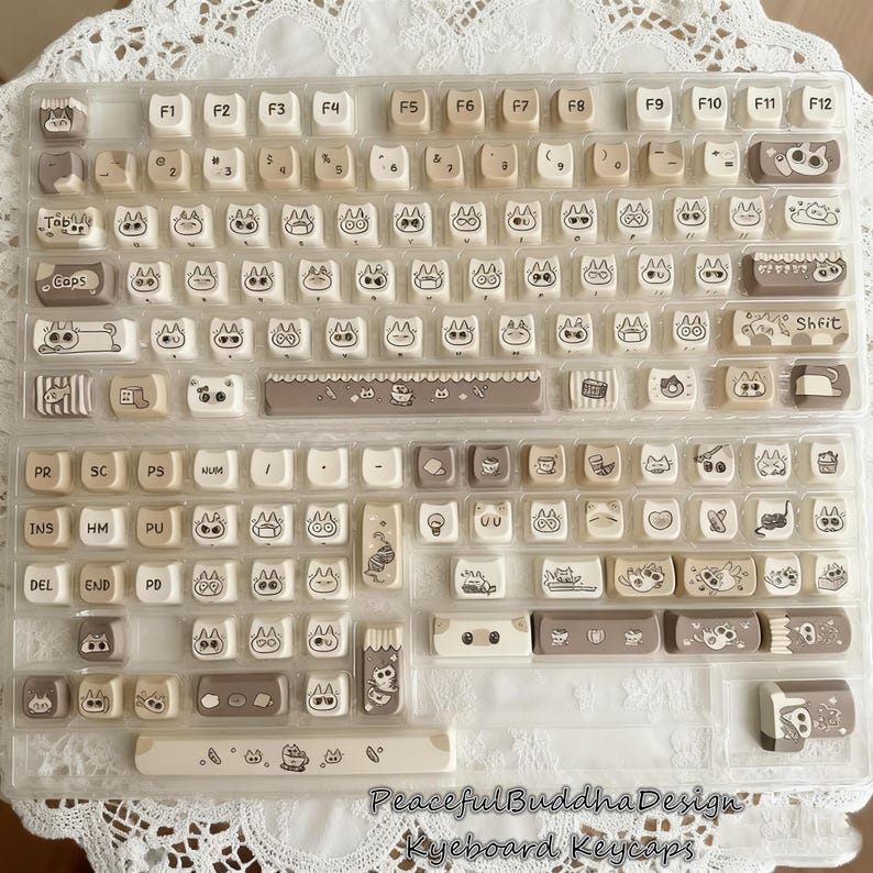 May include: A set of beige and white keyboard keycaps with cat-themed designs. The keycaps feature various cat illustrations and icons, with the text "Peaceful Buddha Design Keyboard Keycaps" at the bottom.