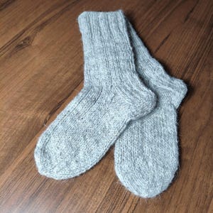 Puede incluir: Un par de calcetines de punto gris claro. Los calcetines están hechos de un hilo suave y texturizado y tienen un puño y pie acanalados. Los calcetines están sobre una superficie de madera.