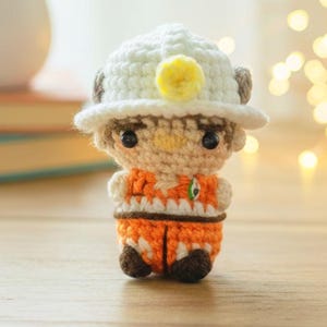 May include: A handmade crochet doll of a miner, wearing an orange and brown outfit and a white hat with a yellow pom-pom. The doll has black eyes and a neutral skin tone. The background includes a blurred succulent plant and a string of lights.