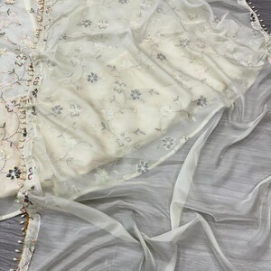 May include: Off-white embroidered fabric with floral patterns and a sheer overlay. The edges are trimmed with a delicate beaded border. The fabric appears to be part of a traditional garment.