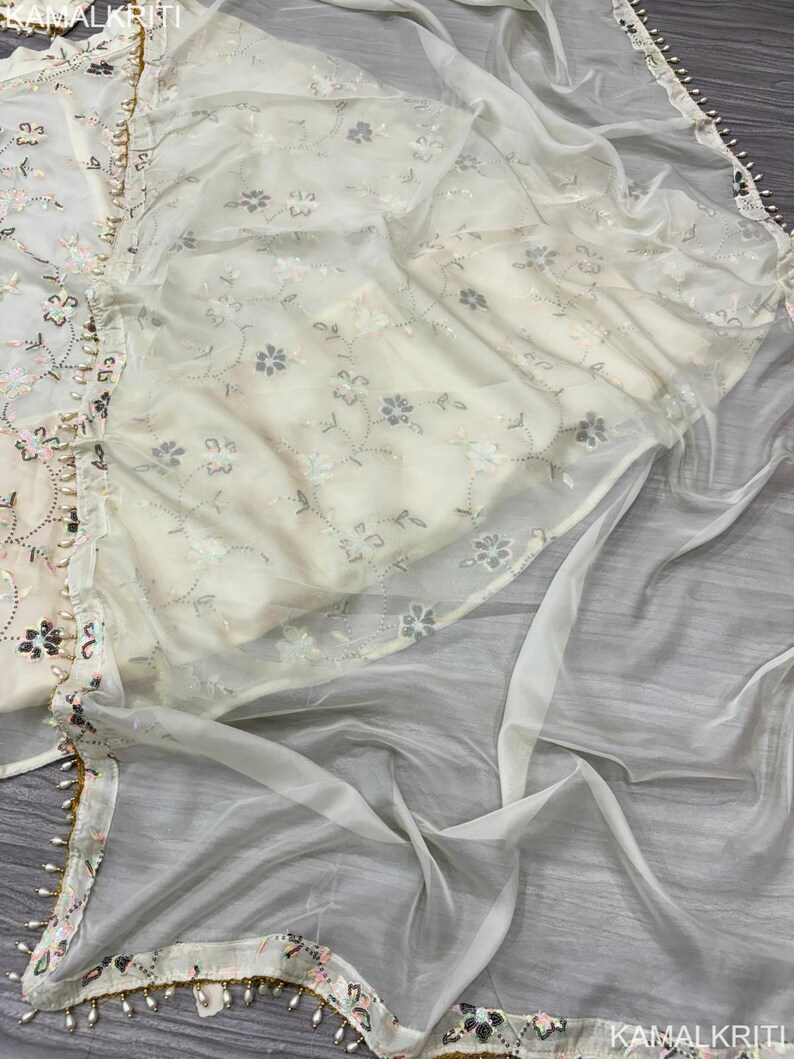 May include: Off-white embroidered fabric with floral patterns and a sheer overlay. The edges are trimmed with a delicate beaded border. The fabric appears to be part of a traditional garment.