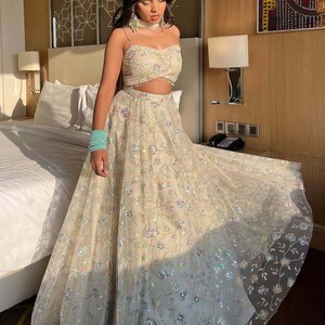 May include: A cream-colored two-piece formal outfit. The top is a crop top with thin straps and floral embroidery. The skirt is a full-length skirt with matching floral embroidery. The outfit is made of a sheer fabric. The text "KAMALKRITI" is visible.