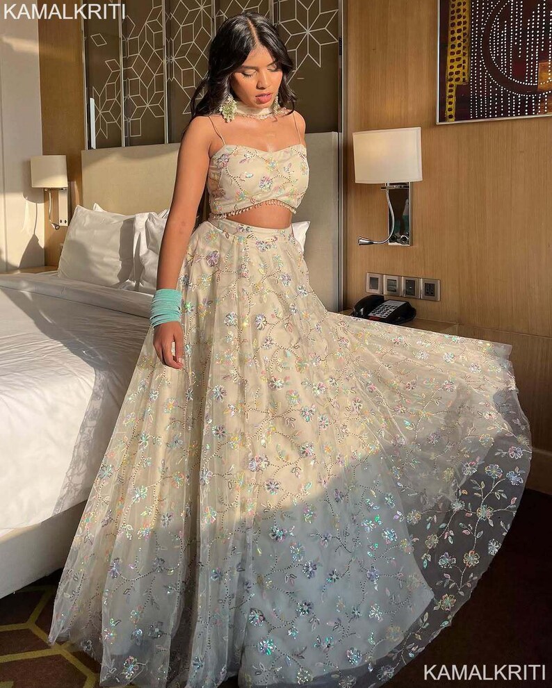 May include: A cream-colored two-piece formal outfit. The top is a crop top with thin straps and floral embroidery. The skirt is a full-length skirt with matching floral embroidery. The outfit is made of a sheer fabric. The text "KAMALKRITI" is visible.