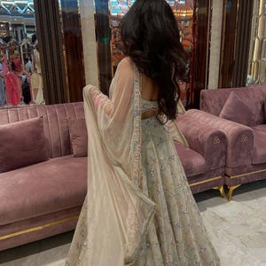 May include: A light-colored, embroidered formal gown with a matching sheer shawl. The gown features intricate floral patterns and embellishments. The background includes a pink velvet sofa and gold accents. The image is branded with the text "KAMALKRITI".