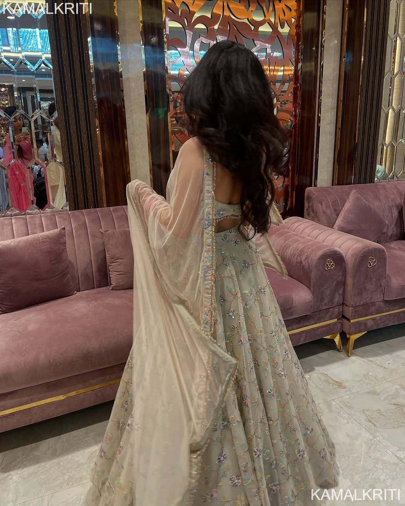 May include: A light-colored, embroidered formal gown with a matching sheer shawl. The gown features intricate floral patterns and embellishments. The background includes a pink velvet sofa and gold accents. The image is branded with the text "KAMALKRITI".
