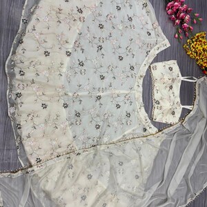 May include: Ivory-colored embroidered formal wear set. The set includes a top, skirt, and sheer scarf, all adorned with delicate floral embroidery in shades of pink and brown. The sheer scarf has a beaded edge. The brand name "KAMALKRITI" is visible.