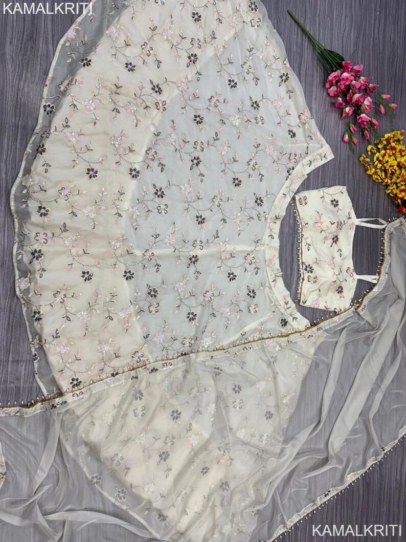 May include: Ivory-colored embroidered formal wear set. The set includes a top, skirt, and sheer scarf, all adorned with delicate floral embroidery in shades of pink and brown. The sheer scarf has a beaded edge. The brand name "KAMALKRITI" is visible.