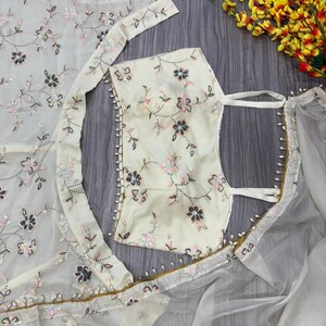 May include: Ivory-colored embroidered fabric with floral patterns. The fabric is adorned with sequins and beads, and is part of a garment. The image also shows a matching top and a sheer overlay, all with delicate floral embroidery.
